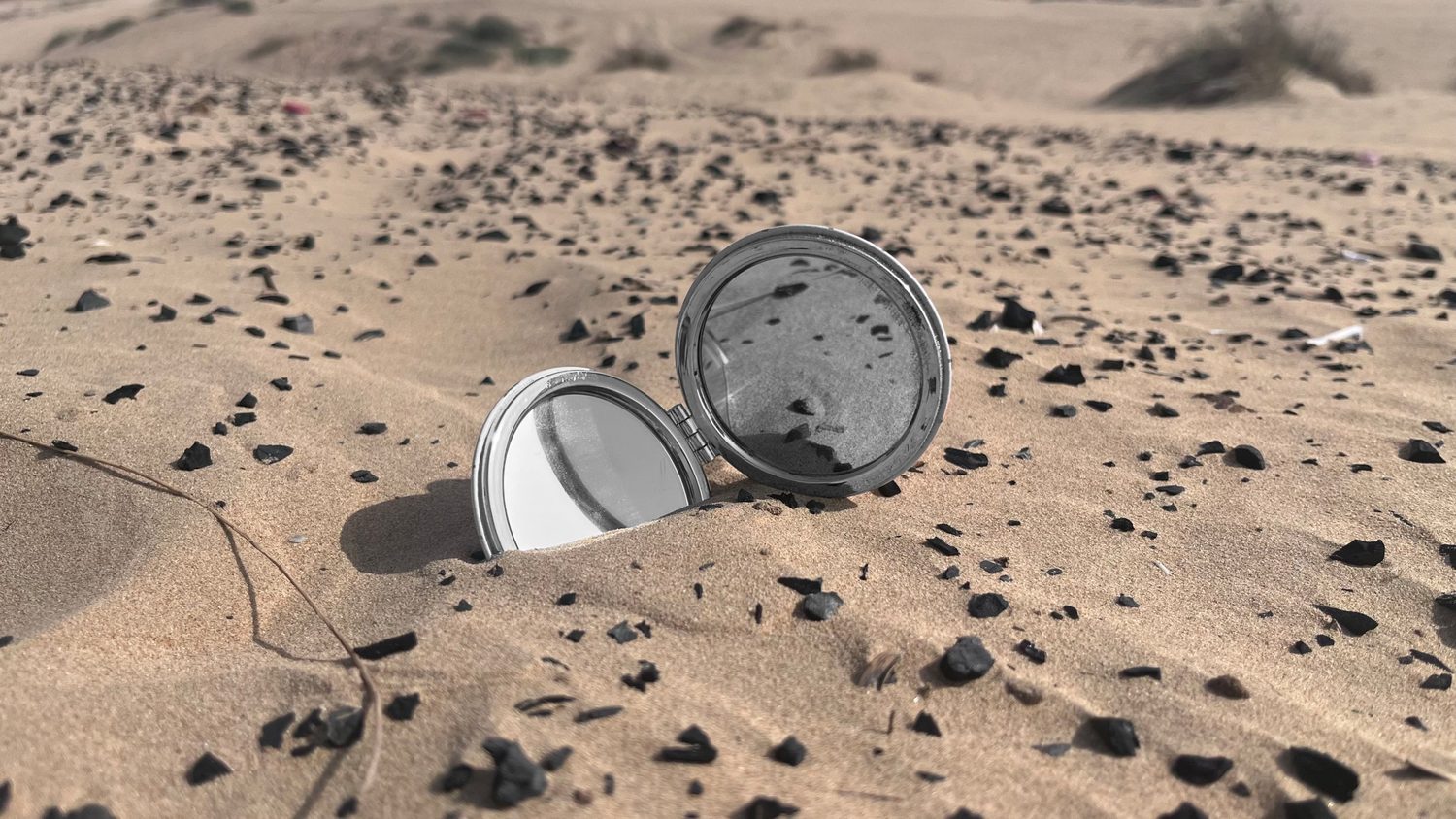 An open compact mirror half-buried in rippled sand scattered with black stones