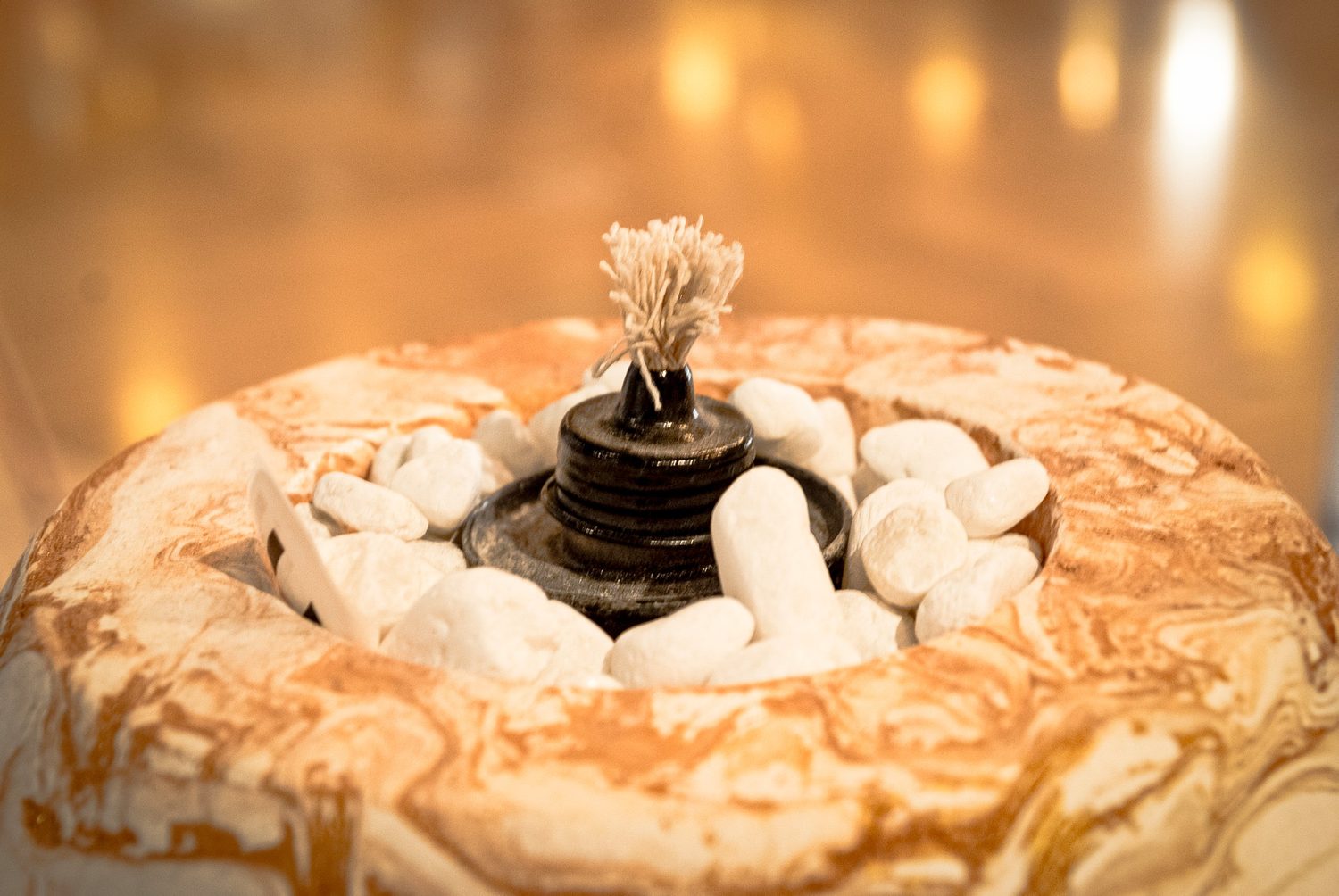 Lit candle set in white pebbles inside a marbled ceramic bowl, warm bokeh behind